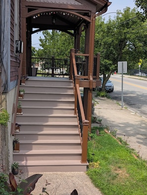 Eine Holztreppe führt zu einer überdachten Veranda mit Geländer, umgeben von Grünpflanzen und mit Blick auf die Straße.