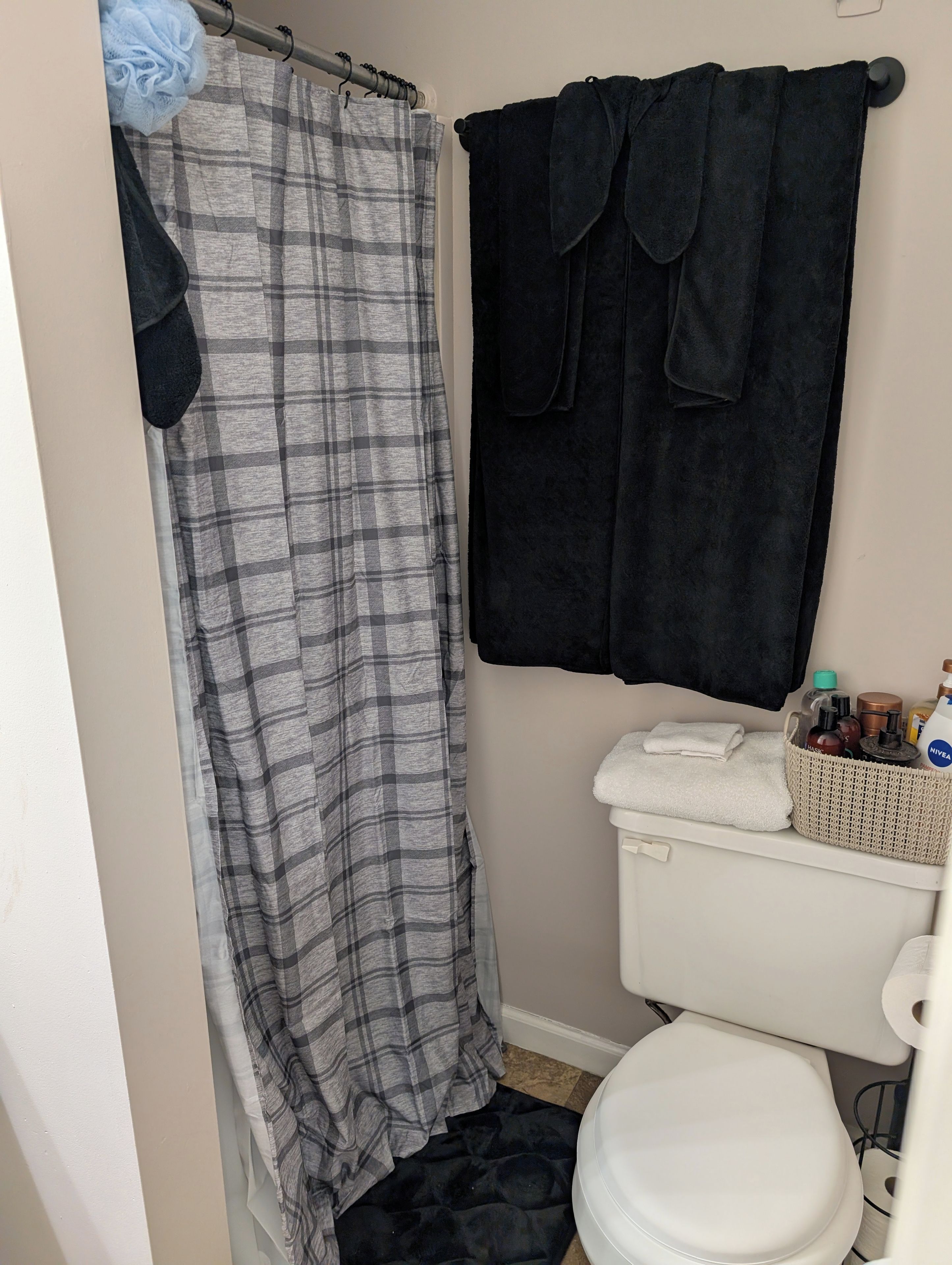 A bathroom with a white toilet, a black towel, a grey plaid shower curtain, and a black bath mat.