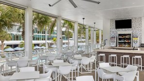 A modern outdoor bar area with white tables and chairs, a pool view, and a bar stocked with various beverages.