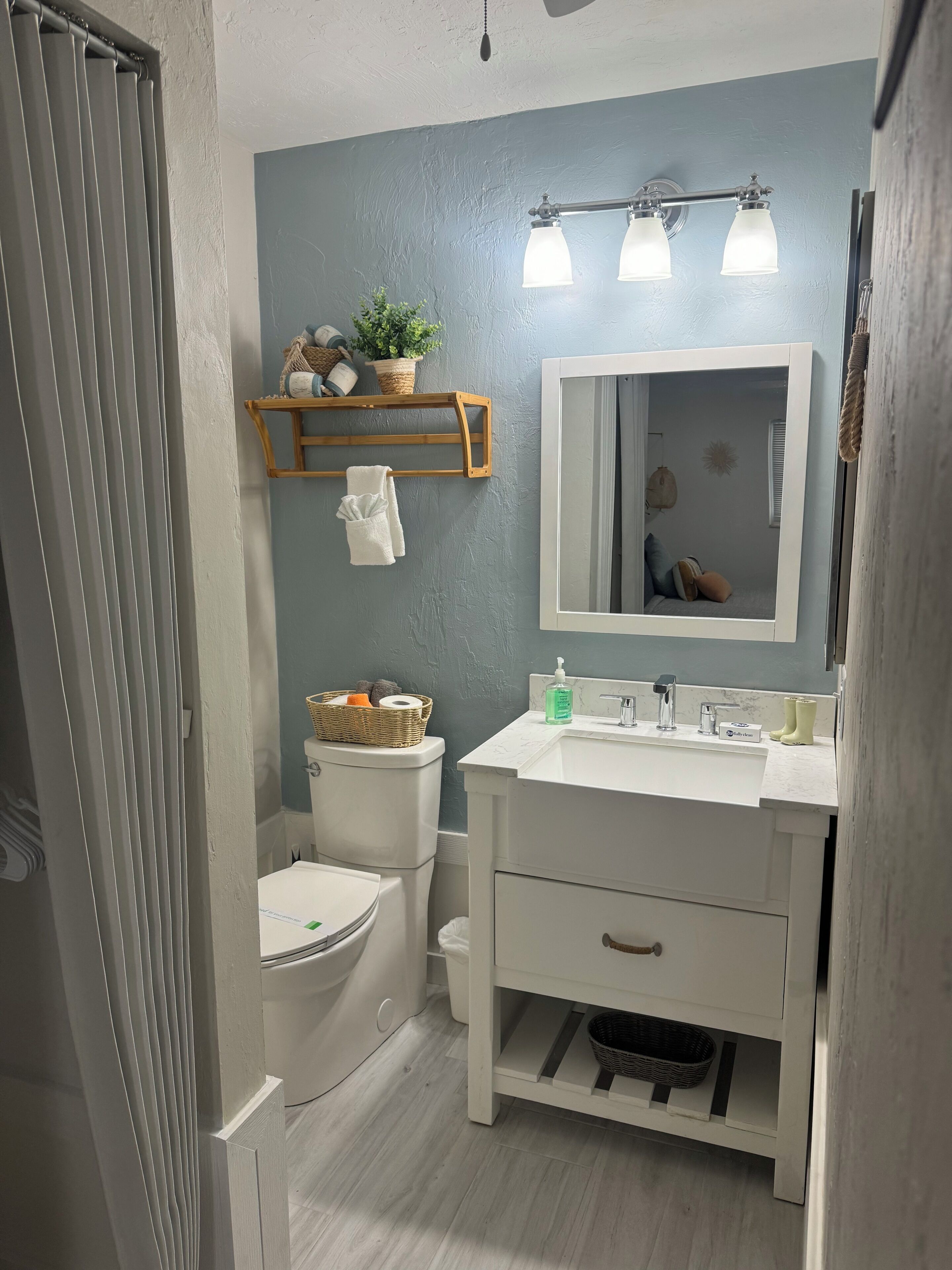 A bathroom with a toilet, sink, and mirror. There is a shelf above the toilet with a plant and a basket.