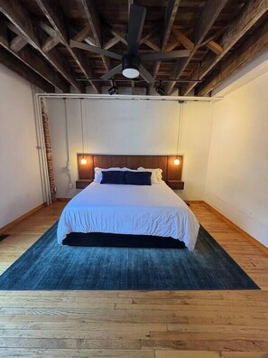 A bedroom with a bed, wooden floor, a ceiling fan, and a brick wall.