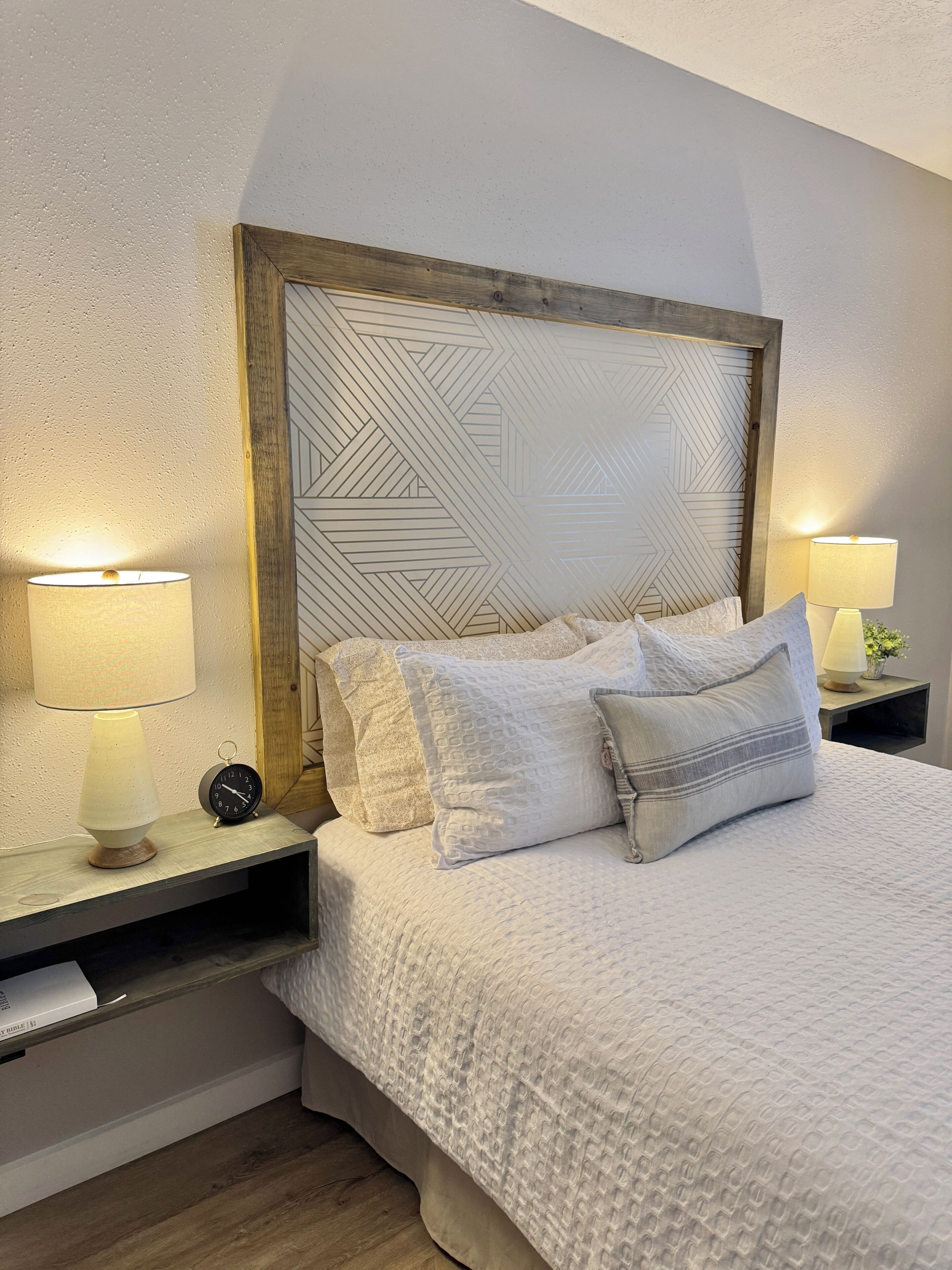 Un lit bien fait, recouvert d’une couette blanche à texture intéressante, deux lampes de chevet, une tête de lit en bois ornée d’un motif géométrique et une petite horloge analogique posée sur la table de chevet.