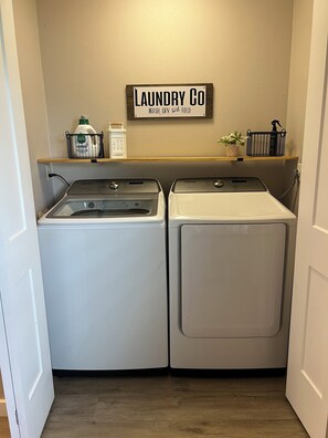 A laundry room with two front-loading washing machines, a shelf with laundry supplies, and a sign above that reads 'Laundry Co Wash, Dry and Fold.' - Rest Haven - Cozy, 2 bedroom Townhome (Spring)