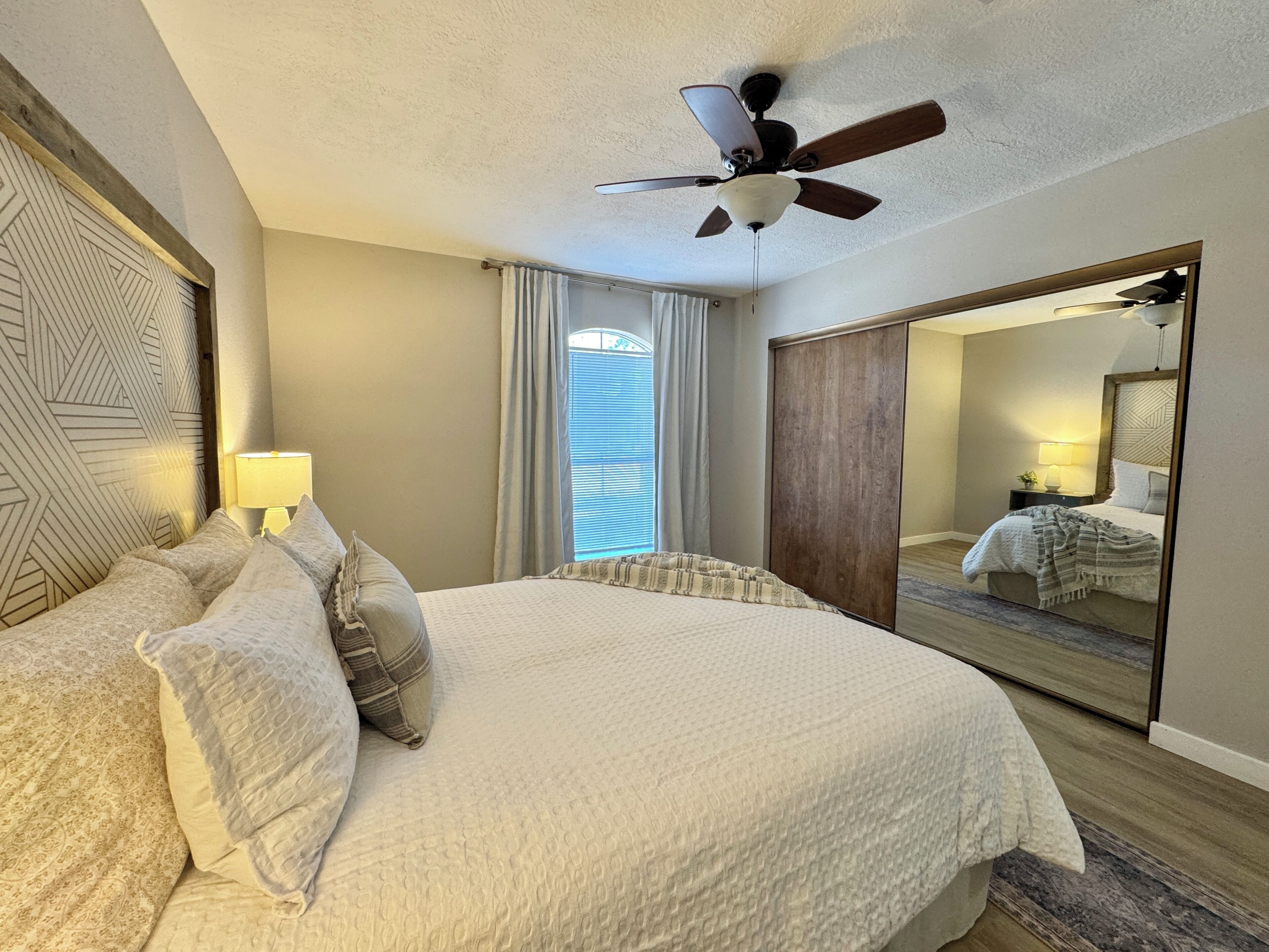 A hotel room with a bed, a ceiling fan, a large mirror, and a window with curtains.