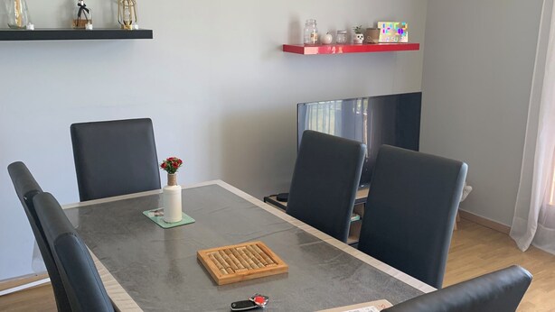 A dining area with a rectangular table, six chairs, a vase with flowers, a chessboard, a remote control, and a box.