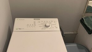 A front-loading white washing machine with a control panel, located in a corner of a room with a white wall and a small window.