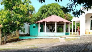 A covered outdoor area with a paved walkway, a small building with a green exterior, and a white building with a red roof in the background. - Girardot Vacation Rental: Your Relaxation Guaranteed (Cundinamarca)