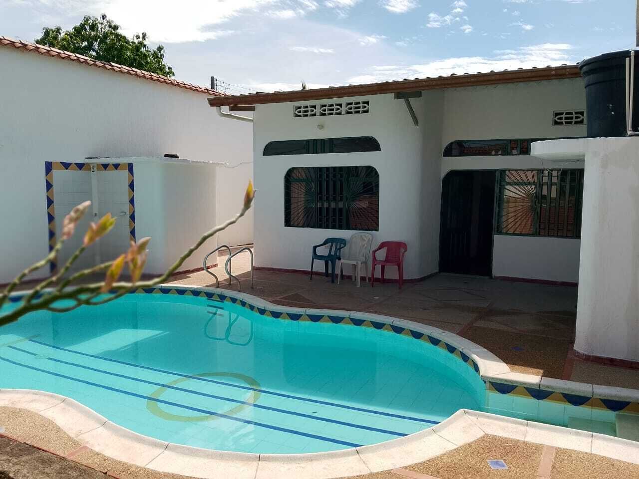 A swimming pool with a tiled edge, a white wall with a window, and a door. There are plastic chairs and a water tank visible.