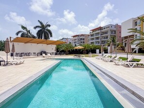 A swimming pool with lounge chairs and umbrellas, surrounded by palm trees and a building with balconies. - Sophisticated Maho Studio with Resort-Style Amenities in a Gated Community. (Maho)
