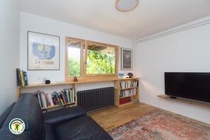 A living room with a black leather sofa, wooden bookshelves, a large flat-screen TV, and a window with a view of greenery. - Le Bi-Fûts - ALLEVARD (ALLEVARD)