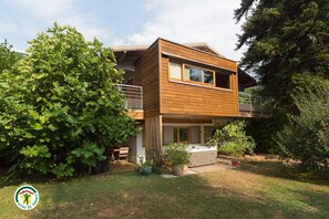 Ein zweistöckiges Haus mit Holzfassade, einem Balkon und einem Garten mit einer großen Feigenbaum.