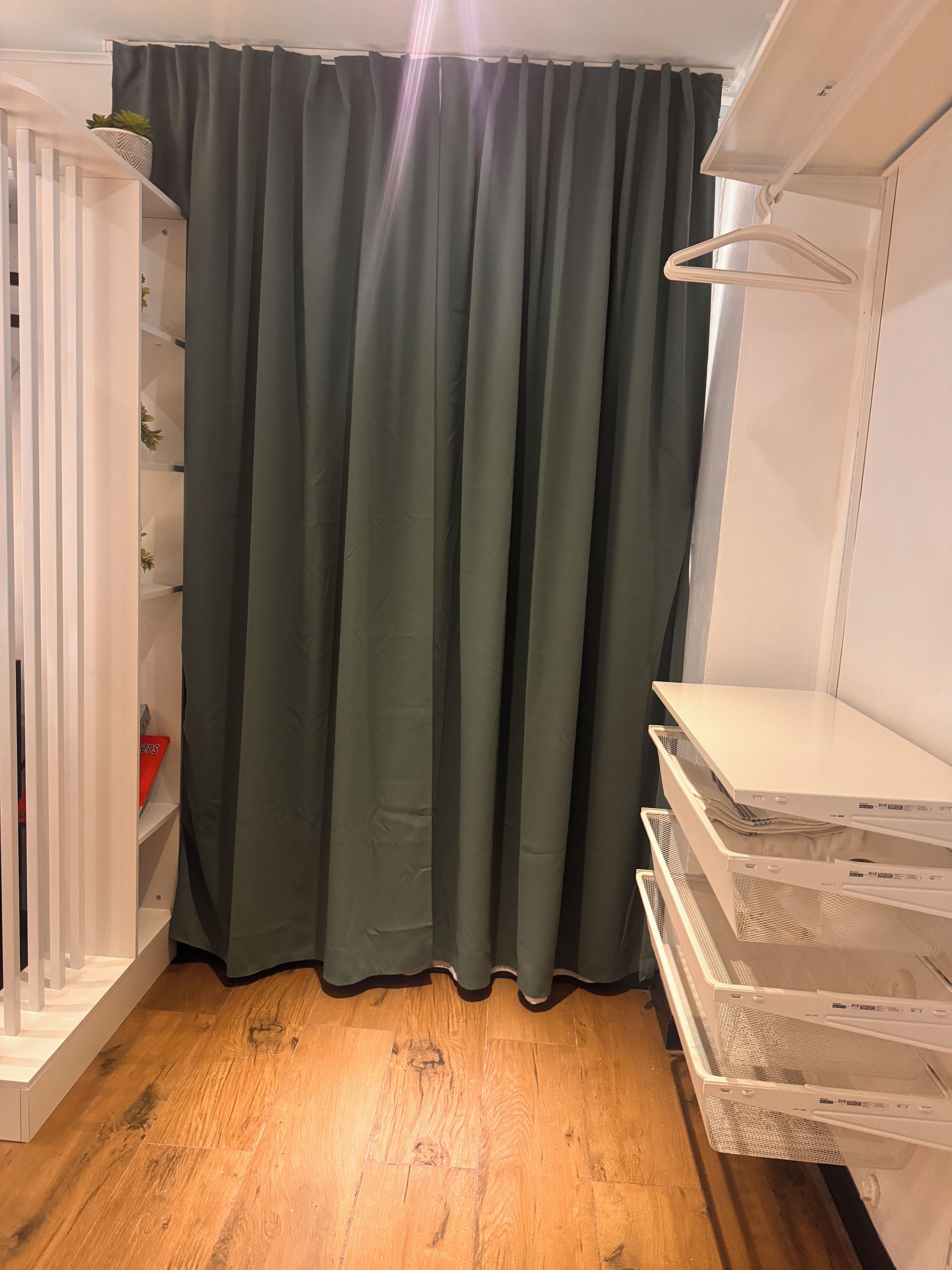 A room with a wooden floor, a green curtain, a white shelf unit, and a white hanging rack.