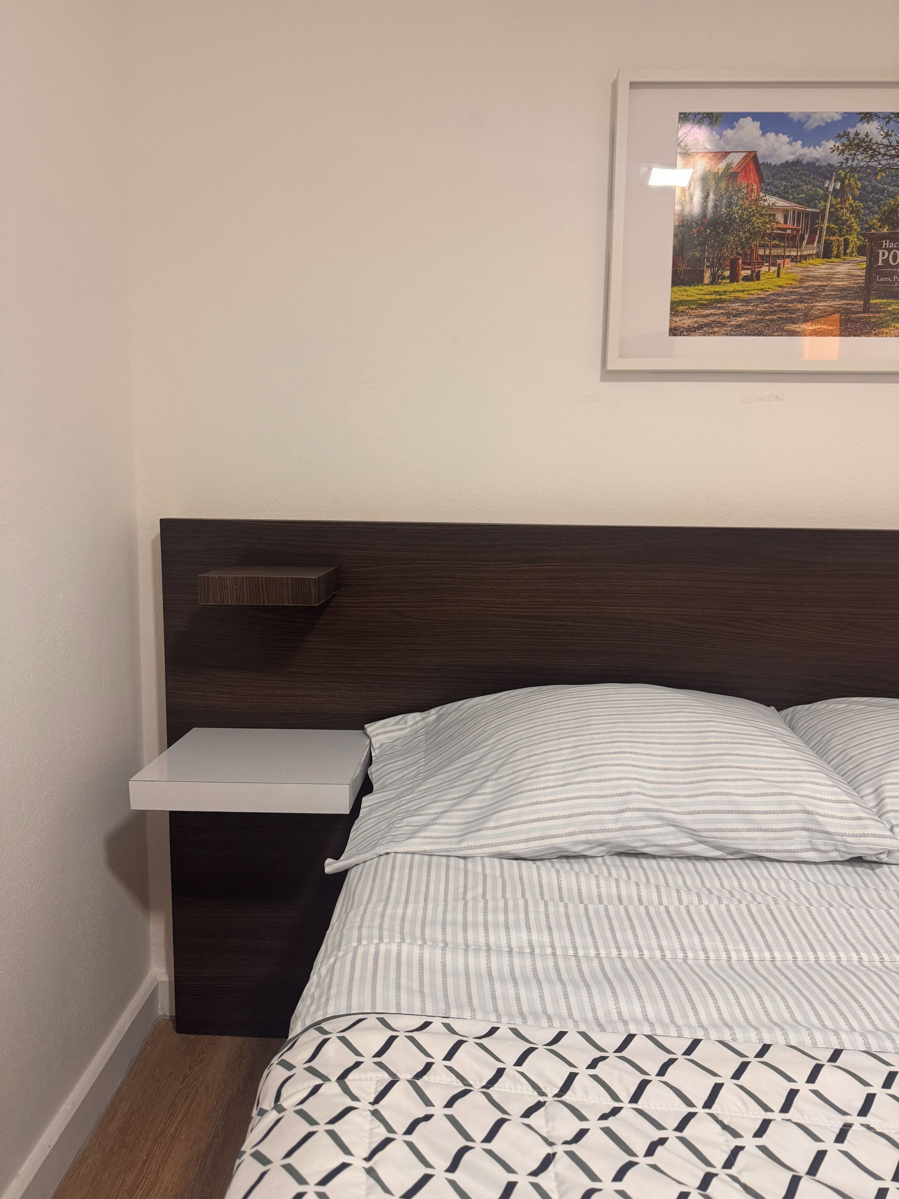 A bedroom with a dark wood headboard, a bedside table, a framed picture on the wall, and a patterned bedspread.
