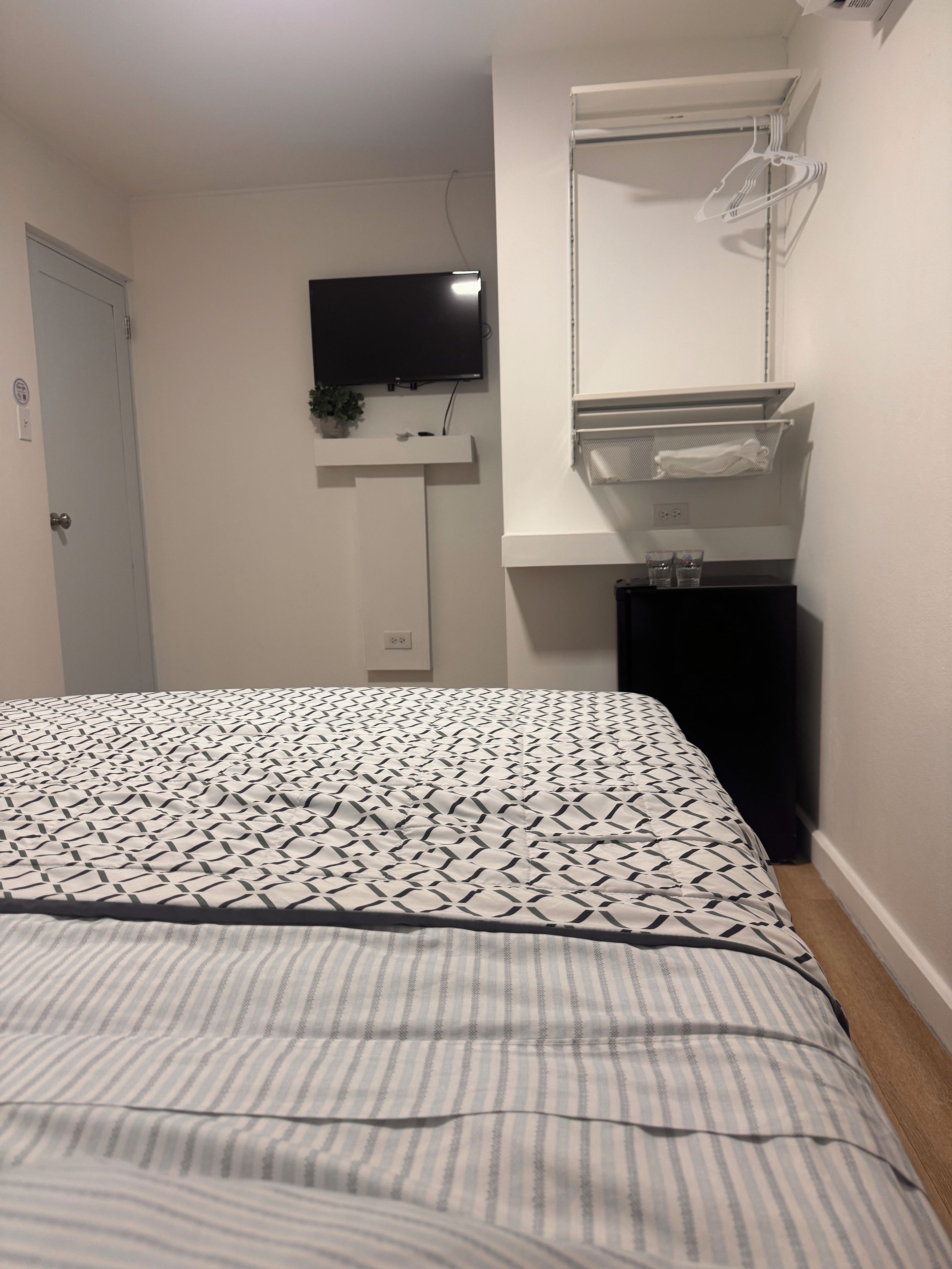 Een slaapkamer met een bed, een aan de muur bevestigde televisie, een klein plankje met een plant en een kledingrek.