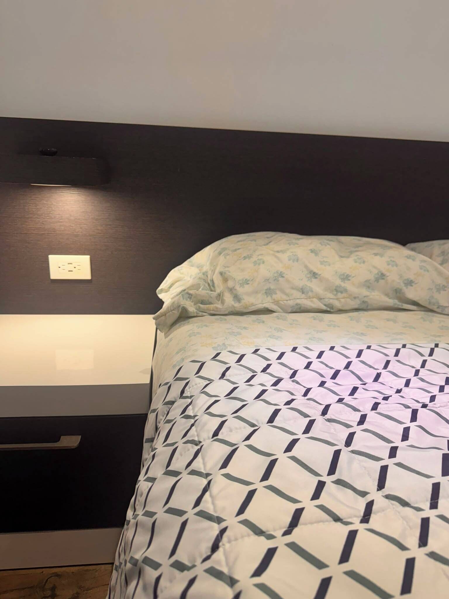 Una cama con una colcha estampada y un cojín floral, una mesita de noche con toma de corriente y un interruptor de luz en la pared.