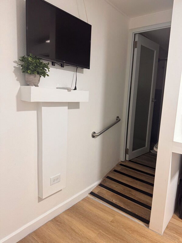 A hallway with a mounted television, a potted plant, a handrail, and a door leading to another room.