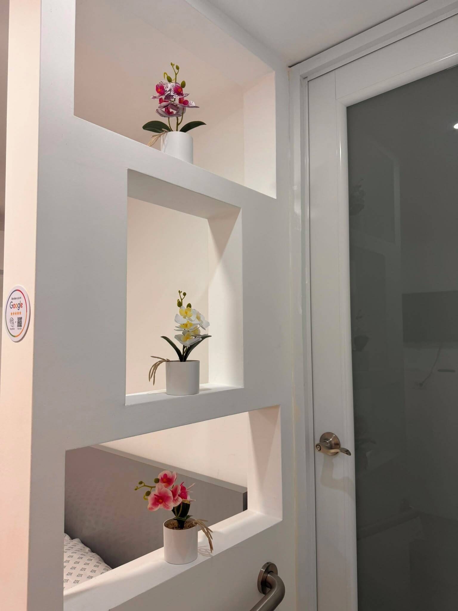 A bathroom with a white door, a glass shower enclosure, and three white shelves with potted orchids.