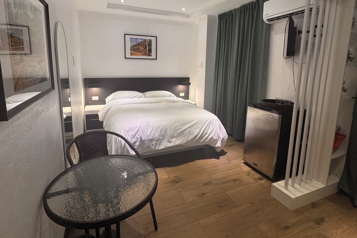 A hotel room with a bed, a small table, a chair, a refrigerator, and an air conditioning unit.