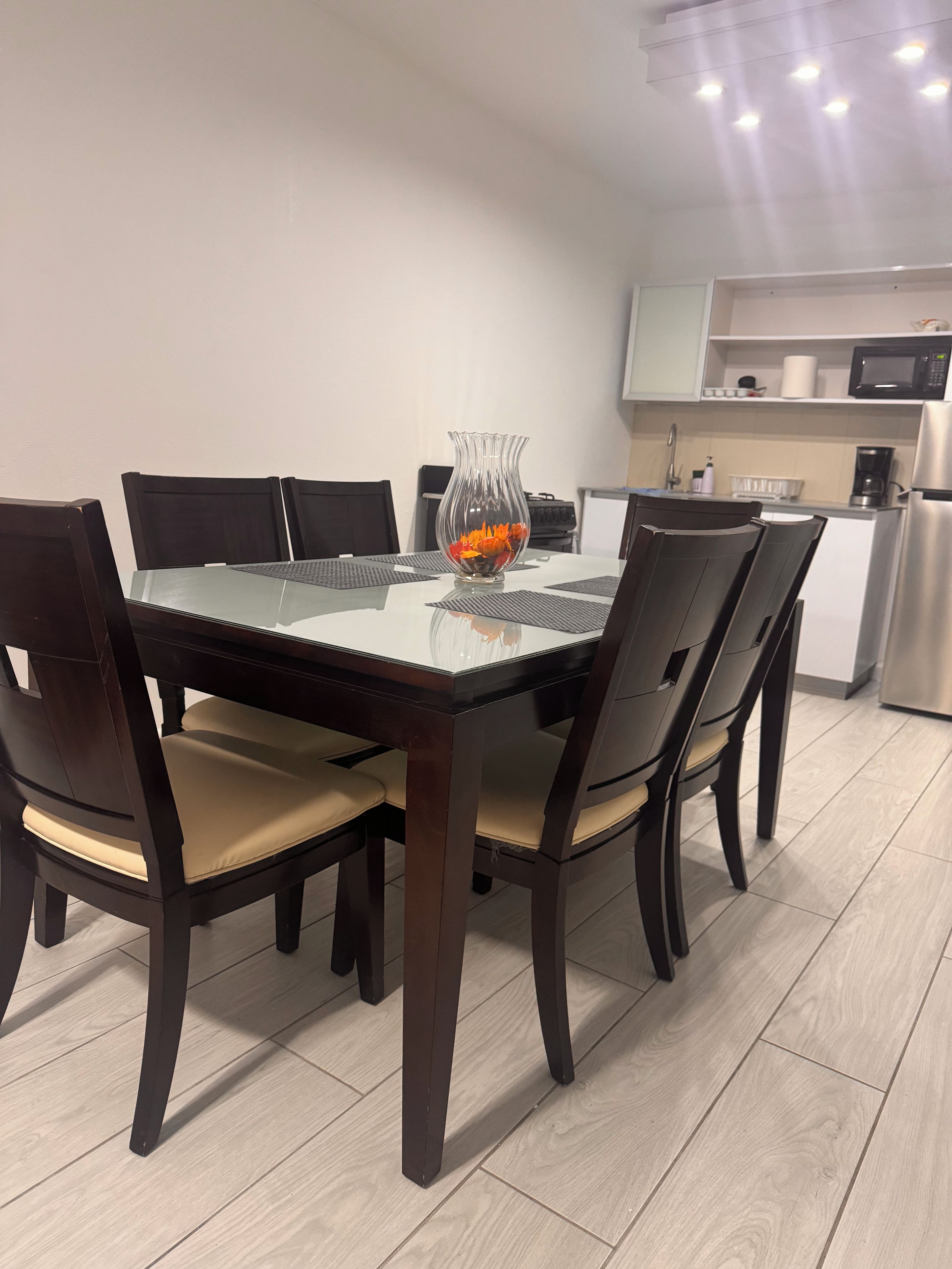 Un comedor con mesa de vidrio, sillas de madera oscura y un florero con flores naranjas.