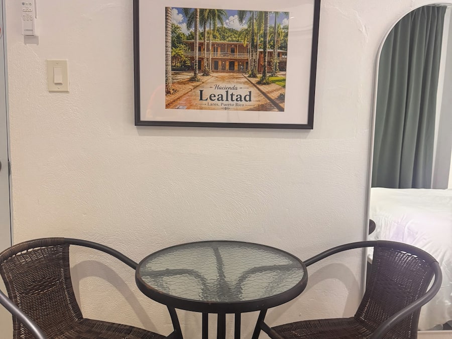 Un cuadro enmarco muestra un edificio con el texto 'Hacienda Lealtad Lares, Puerto Rico' en una pared. Frente a él, hay una mesa redonda de vidrio y dos sillas de mimbre.