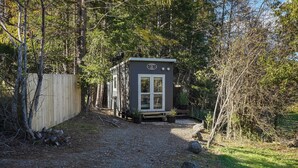 A small, single-story cabin with a door and windows, surrounded by a gravel path and dense trees. - Quiet and Private Tiny Home with Outdoor Bath in Hanmer Springs (Hanmer Springs)