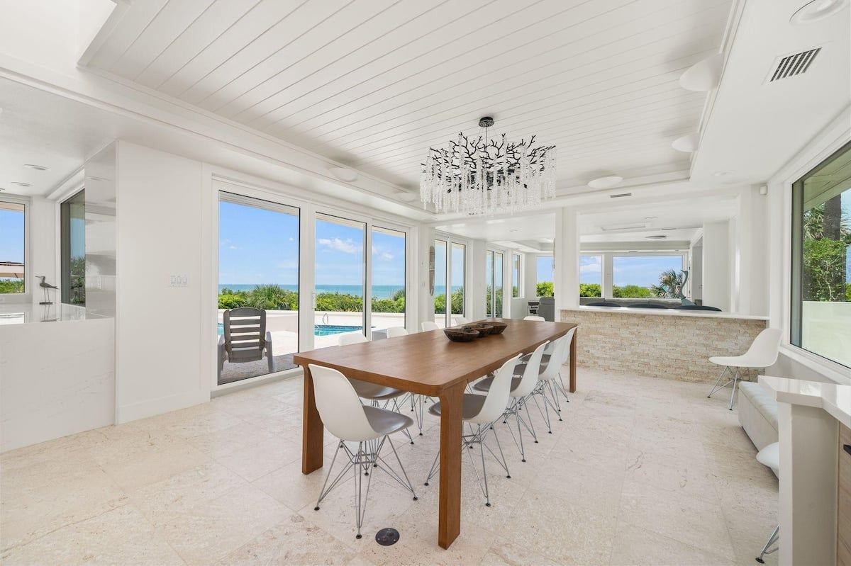 A spacious dining area with a long wooden table and modern chairs, large windows offering a view of the outdoors, and a decorative chandelier.
