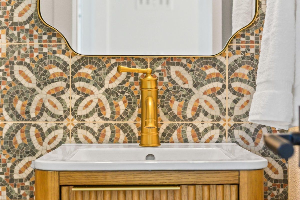 Un baño con salpicadero de mosaicos, grifo dorado y lavamanos blanco.