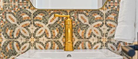 Un baño con salpicadero de mosaicos, grifo dorado y lavamanos blanco.