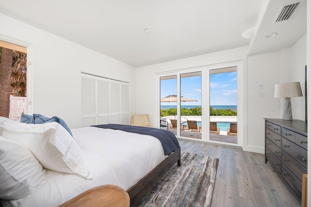 A bedroom with a bed, a dresser, a window with a view of the ocean, and a sliding glass door leading to an outdoor area.