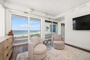 A modern living room with a large window offering a view of the ocean, two armchairs, a flat-screen TV mounted on the wall, and a wooden floor. - Panoramic Paradise I Luxury Beachfront Estate & Boardwalk to the Beach (Saint Augustine)