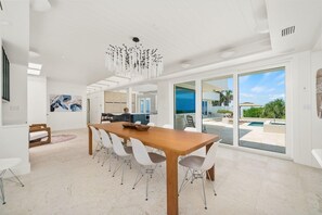 A spacious dining area with a long wooden table and white chairs, a modern chandelier, and a view of a swimming pool through large sliding glass doors. - Panoramic Paradise I Luxury Beachfront Estate & Boardwalk to the Beach (Saint Augustine)