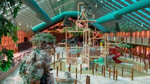 Ein großer Indoor-Wasserpark mit einer zentralen Spielanlage, künstlichen Felsformationen und einer Glasschutzwand.