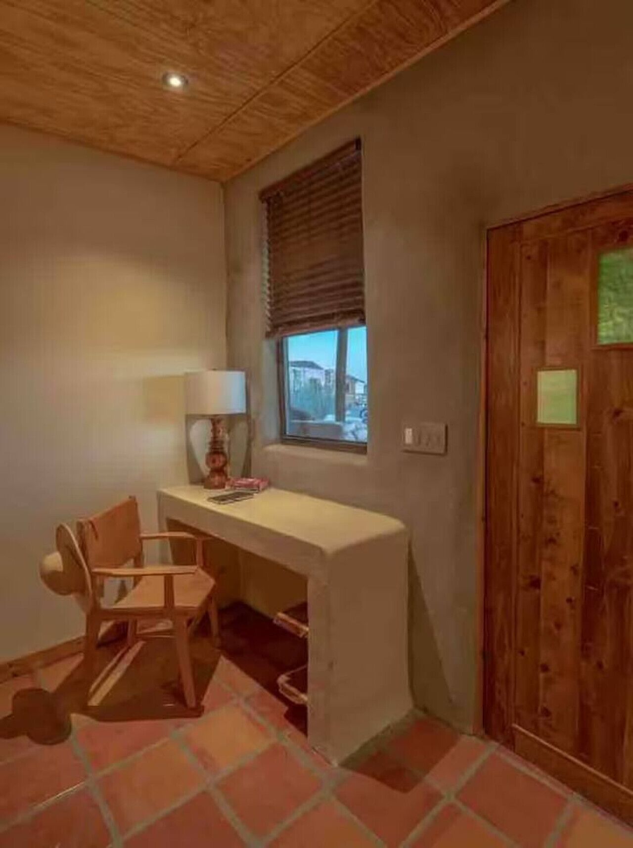 Una habitación con techo de madera, un escritorio con silla, una ventana con persianas y una puerta con un letrero.