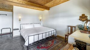 Een slaapkamer met een metalen bedframe, witte beddengoed, een houten plafond, een klein tafeltje met een ventilator en een vloerkleed.