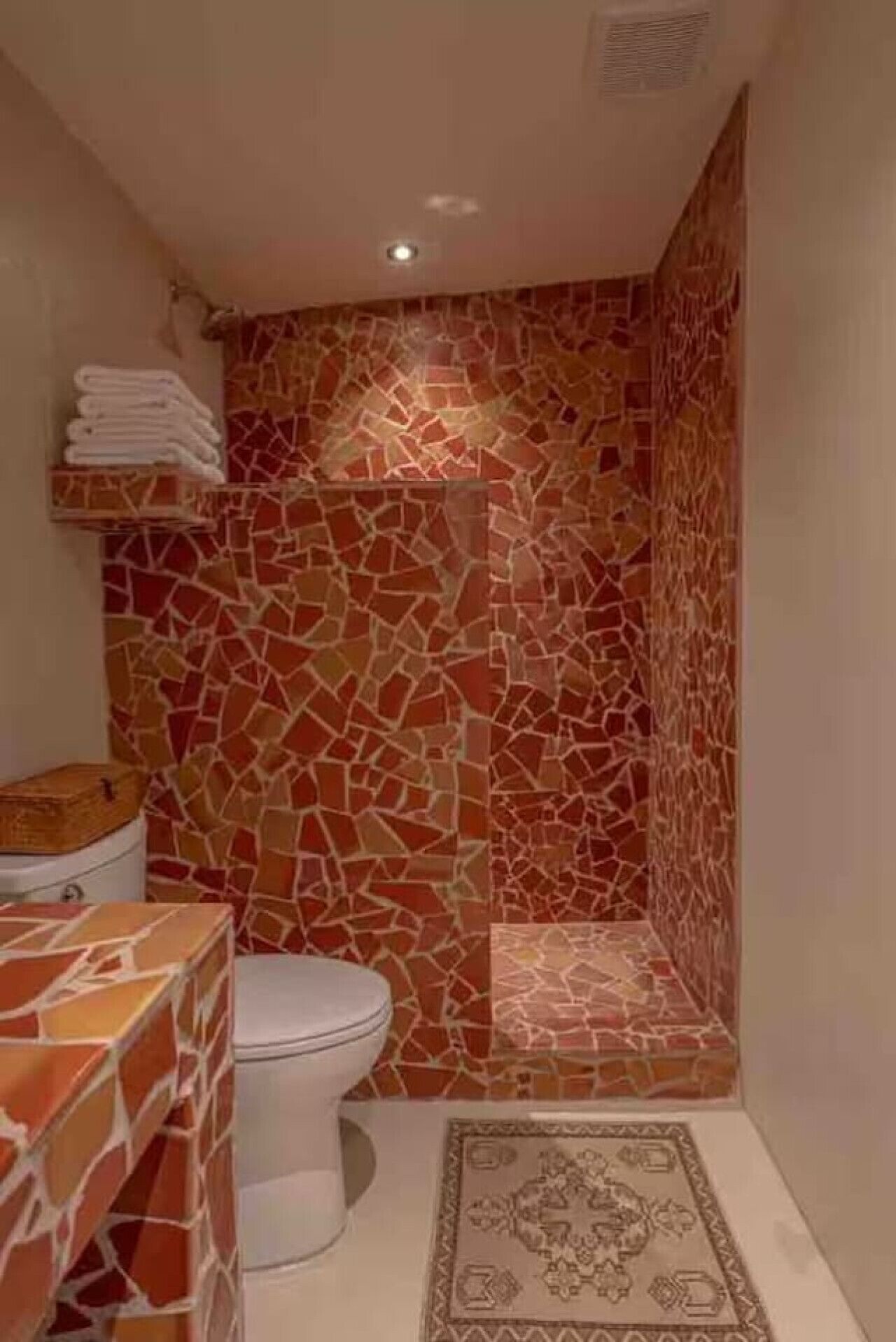 Une salle de bain avec un mur de douche recouvert de mosaïques, des toilettes et un lavabo doté d’un couvercle en bois.