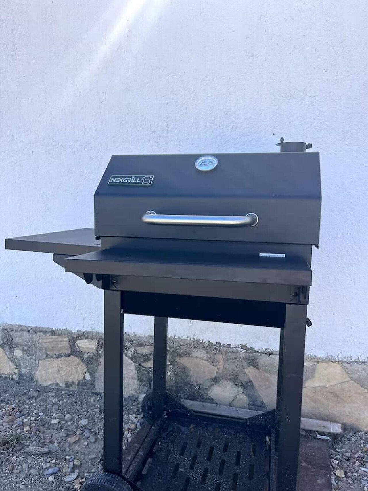 Una parrilla portátil con asa y un logotipo, sobre un soporte metálico, con una pared de piedra al fondo.