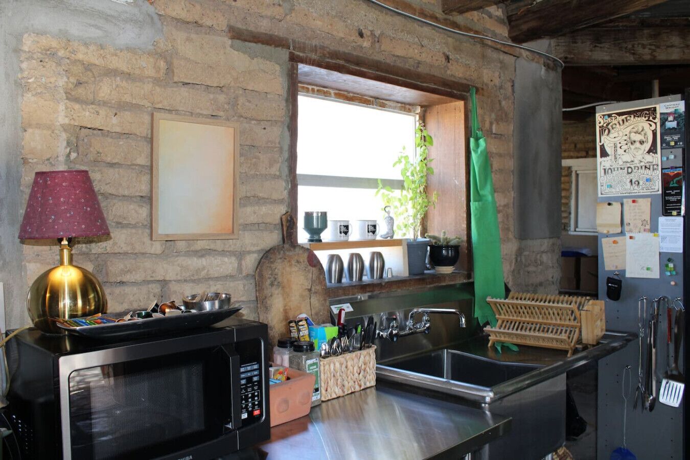 A kitchen with a microwave, sink, and various utensils. There is a window with a view of the outside, and a green apron hanging on the wall.