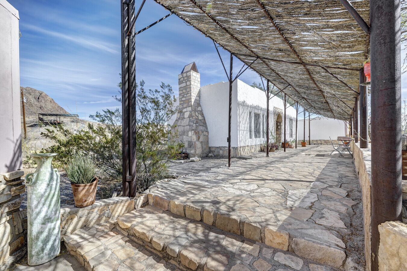 Une cour dallée de pierres, avec des marches menant à un bâtiment blanc, une tour de pierre et un espace ombragé agrémenté d’une pergola.