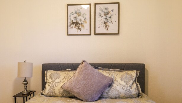A bed with a patterned bedspread, a lamp, and two framed floral artworks on the wall. - Cozy Case Mena . Pet Friendly . Fiber WiFi (San Juan)