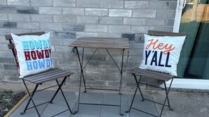 Two metal folding chairs with cushions, one with 'Howdy Howdy Howdy' and the other with 'Hey Y'all', placed on a paved patio against a brick wall. - Cozy Case Mena . Pet Friendly . Fiber WiFi (San Juan)