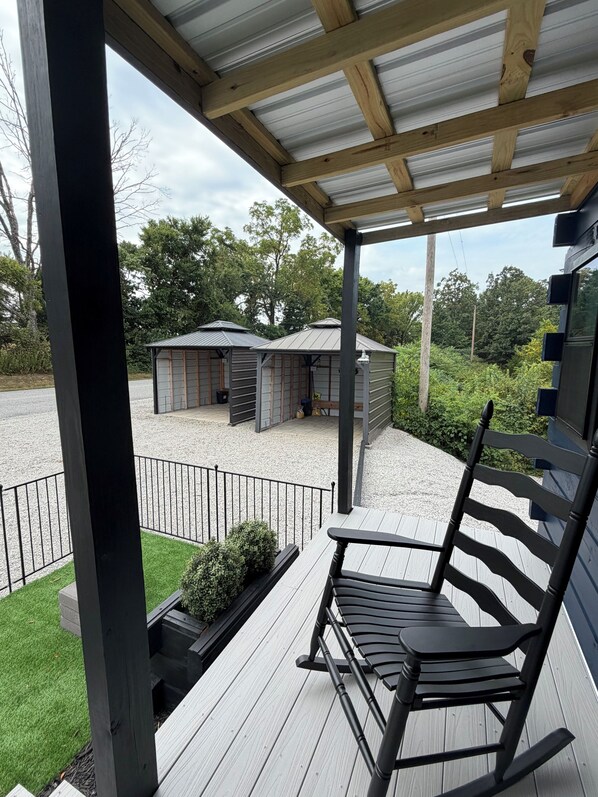 A covered outdoor deck with a wooden chair, a gravel area with a shed, and a view of trees. - Jazzy Moon Log Cabin Gorgeous Ozark Mountain Country View No Cleaning Fees (Eureka Springs)