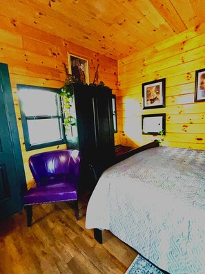 Ein Zimmer mit Holzwänden, einem Bett mit gemustertem Überwurf, einem violetten Sessel, einem dunklen Schrank und gerahmten Bildern an der Wand.