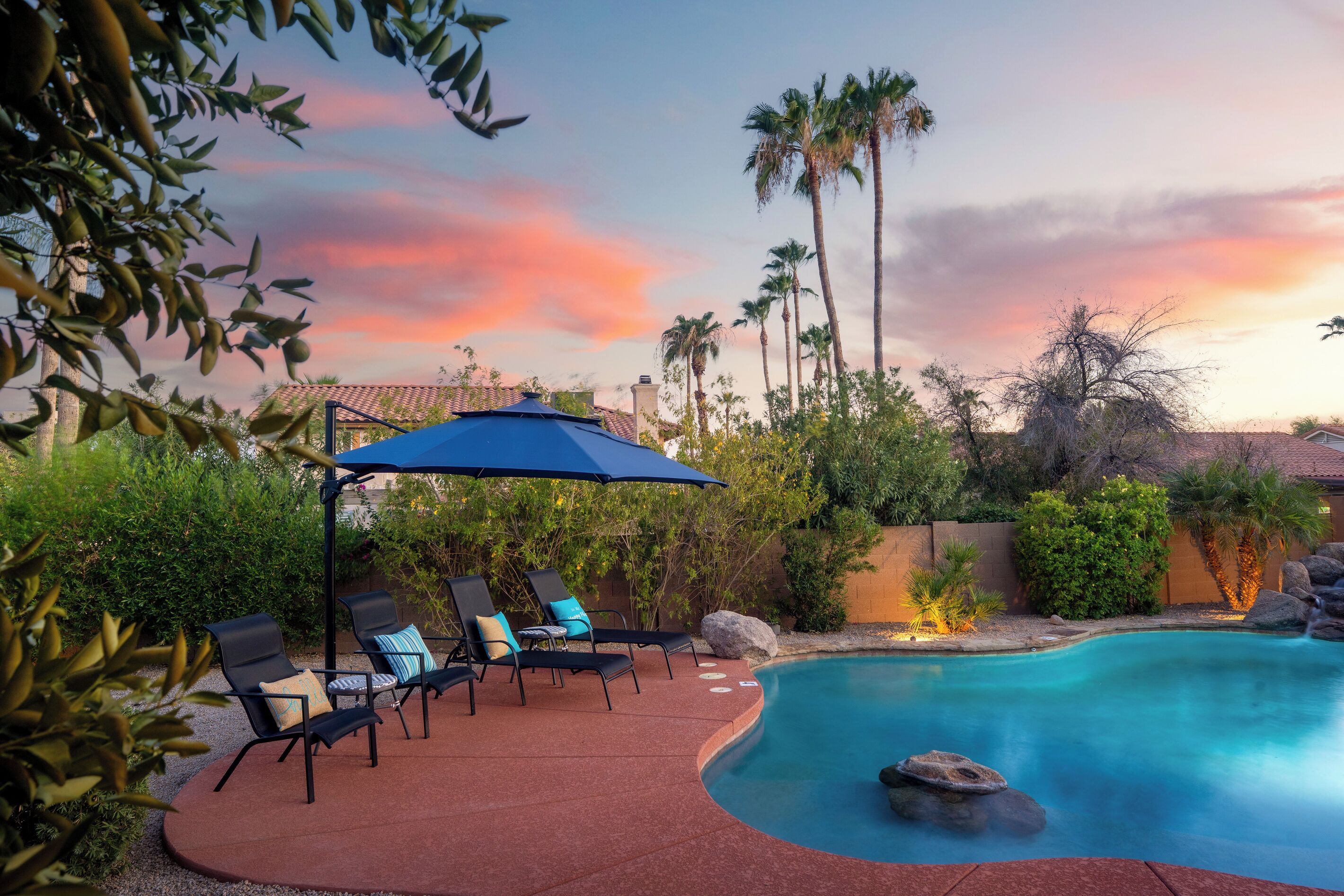 A poolside area with lounge chairs and a covered seating area, surrounded by palm trees and a sunset sky.