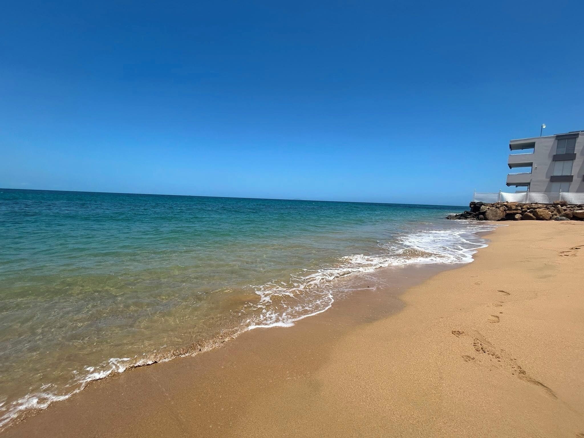 Ein Strand mit klarem Wasser, einem Sandufer und einem Gebäude mit mehreren Balkonen.