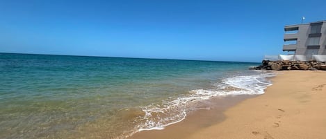 A beach with clear water, a sandy shore, and a building with multiple balconies.