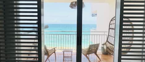 A balcony with two wicker chairs and a small table, overlooking a beach and the ocean.