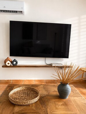 A flat-screen TV mounted on a wall, a wooden shelf with decorative items, a woven basket, and a vase with dried plants on a wooden floor. - Ocean View 2-bedroom 2- bathroom apartment with AC in Rincon (Rincon)