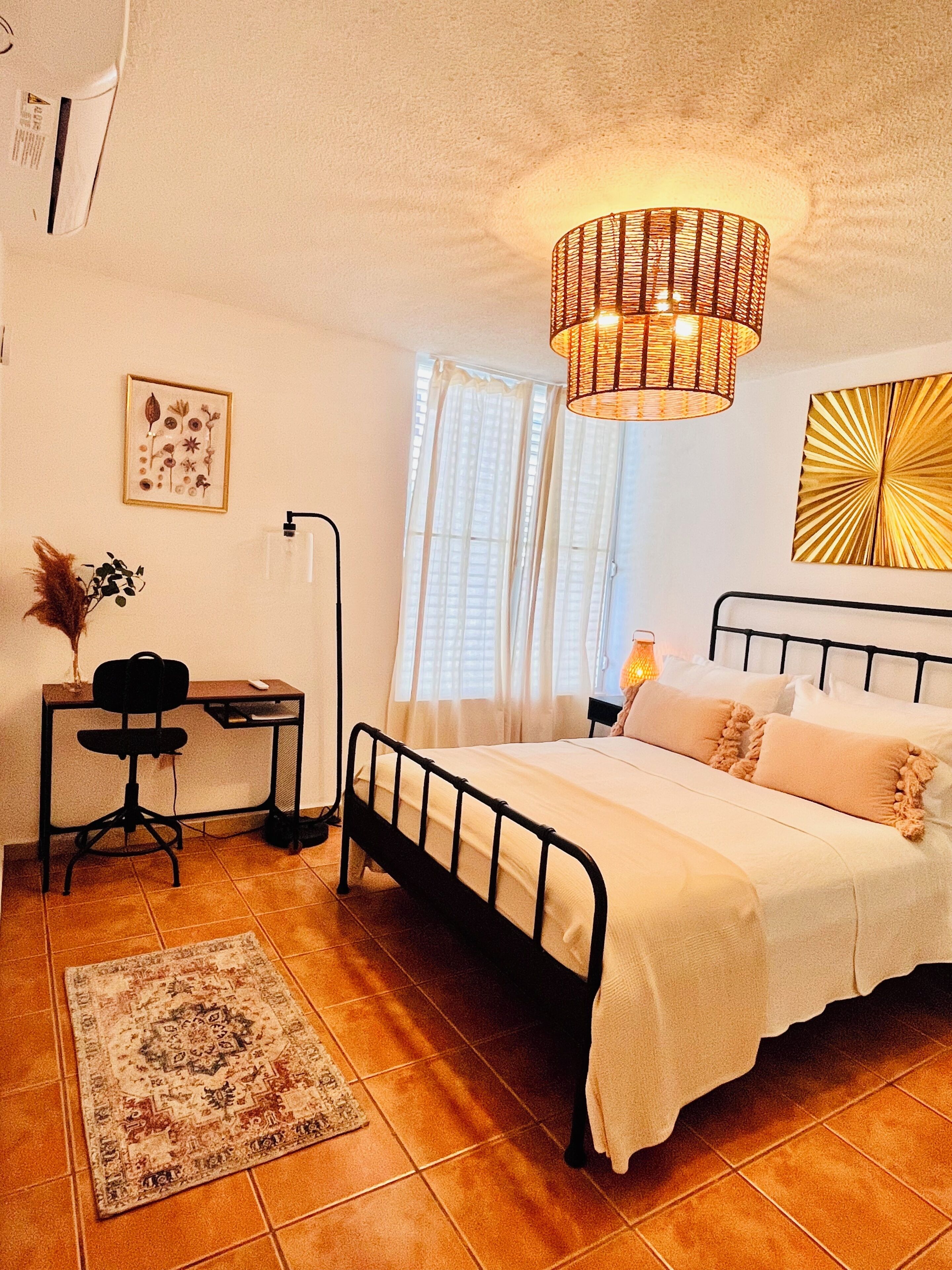 Un dormitorio con cama, escritorio, silla, alfombra, lámpara de pared y ventana con cortinas.