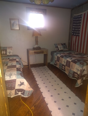 Ein Zimmer mit zwei Betten, einem Nachttisch, einer Lampe, einem Fenster und einer amerikanischen Flagge an der Wand.