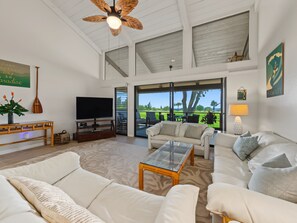 A spacious living area with a television, comfortable seating, a ceiling fan, and a view of a golf course through large windows. - Sunset, Ocean, and Golf Course Views! (Waikoloa Village)